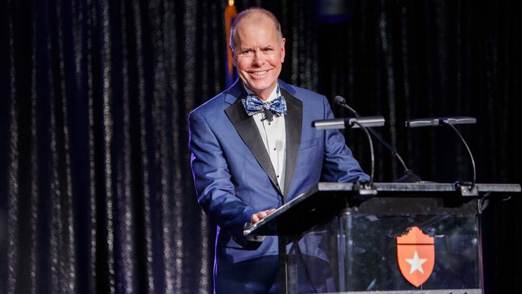 man in blue tuxedo stands behind a podium in front of a black curatin backdrop